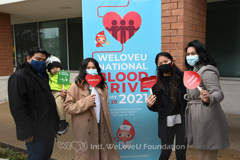 A group of participants take a photo outside with the National Blood Drive standing banner and hold photo props that say 'Donate blood' and 'I saved 3 lives'