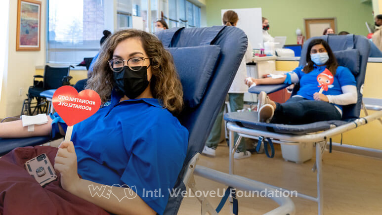 Two blood donors sitting behind each other smile for a photo while donating blood