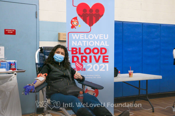 A volunteer is donating blood, smiling and holding up a sign that says Donate Blood, Donate Love.
